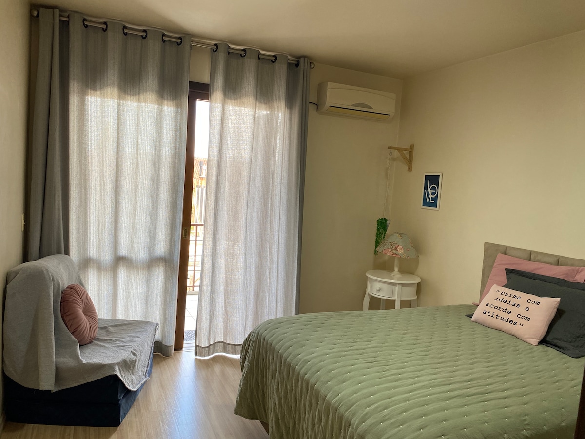 A well-lit bedroom features a spacious bed with a light green coverlet, complemented by a decorative pillow. A comfortable seating area is provided with a gray sofa. Sheer curtains gently filter sunlight, and a small table stands beside the bed, enhancing the room's functionality.