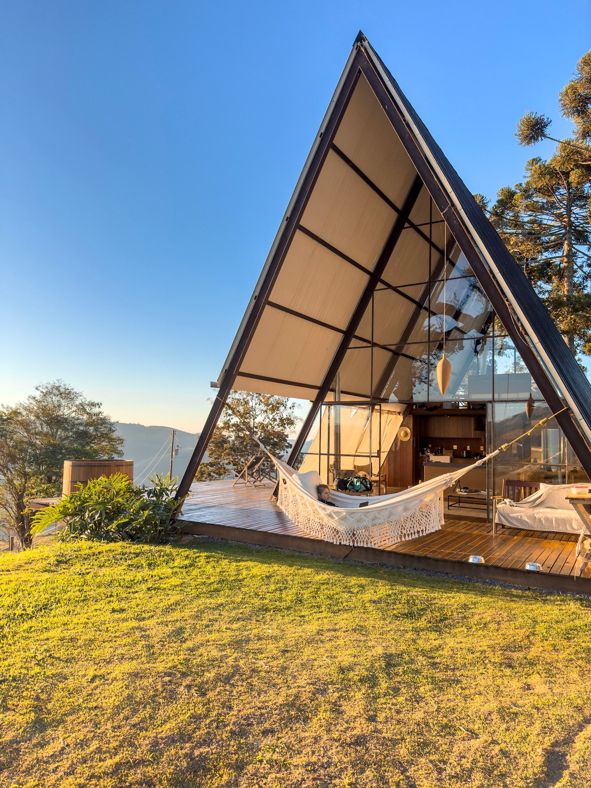 The contemporary cabin features a striking triangular structure with large glass windows. A spacious deck is visible, showcasing two hammocks, set against a backdrop of lush greenery and distant mountains, under a clear, blue sky.