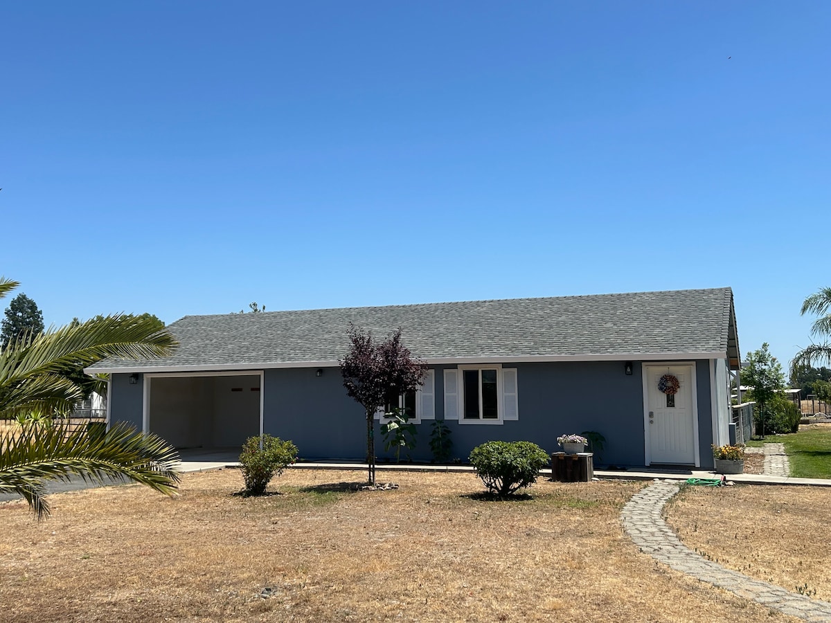 A single-story home presents a neutral blue exterior, complemented by a welcoming front door and windows. Green landscaping surrounds the property, with a small tree and decorative stones leading to the entrance. The driveway offers ample space for parking.