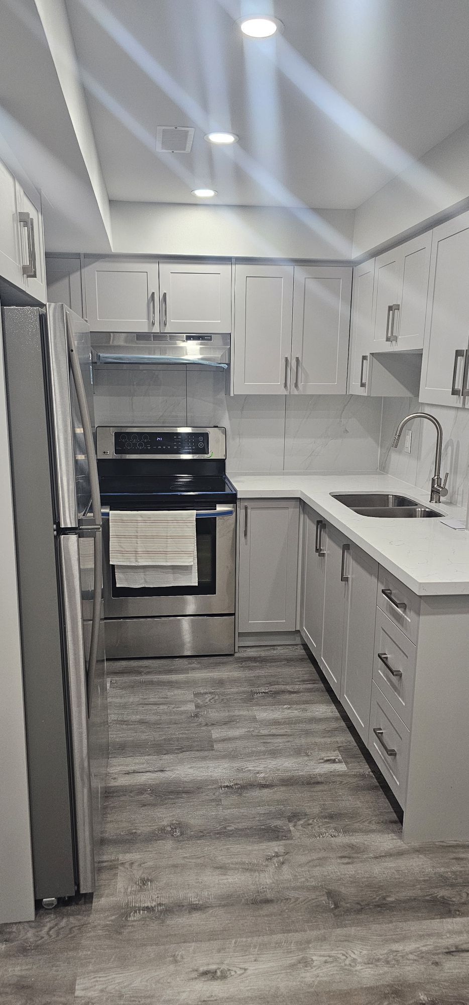 A modern kitchen features stainless steel appliances, including a refrigerator and oven. Sleek cabinetry in light tones lines the walls, with a clean countertop displaying a sink and a dish towel. Bright overhead lighting enhances the contemporary design.