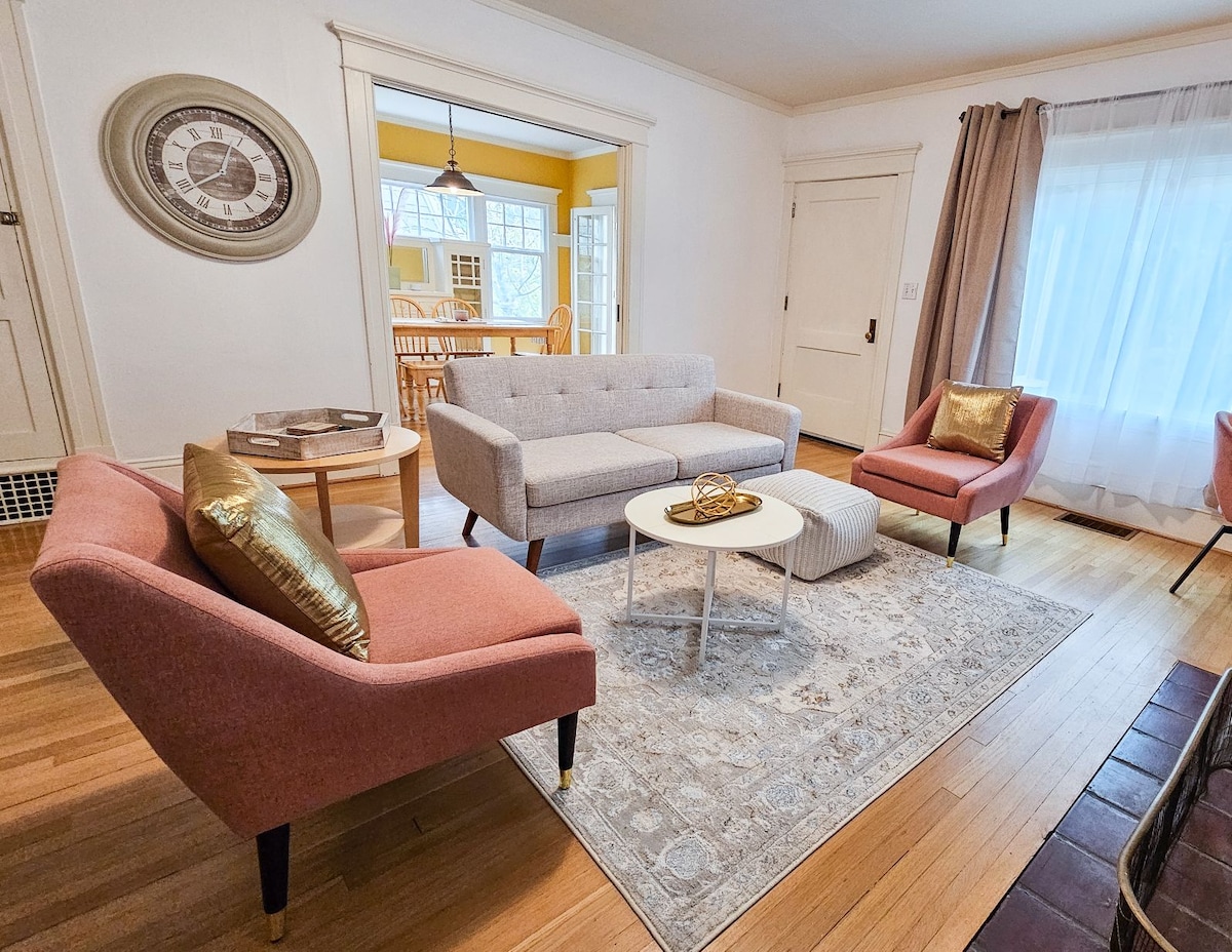 A light-filled living room features a grey sofa and two pink chairs with gold accents. A round white coffee table sits on a textured area rug, complemented by a wall clock and large window draped with sheer curtains, allowing natural light to brighten the space.