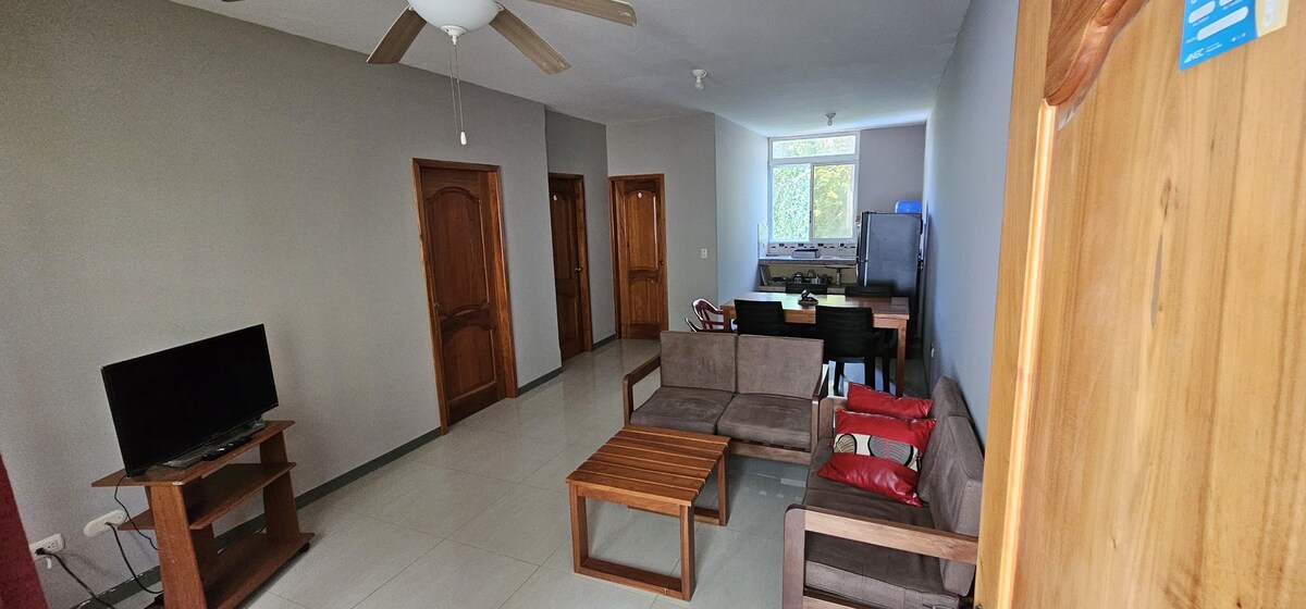 A living area is presented with a comfortable seating arrangement, consisting of two armchairs and a sofa. A wooden coffee table is centrally located, complementing the space. Natural light enters through a window, while a nearby kitchenette is subtly visible.