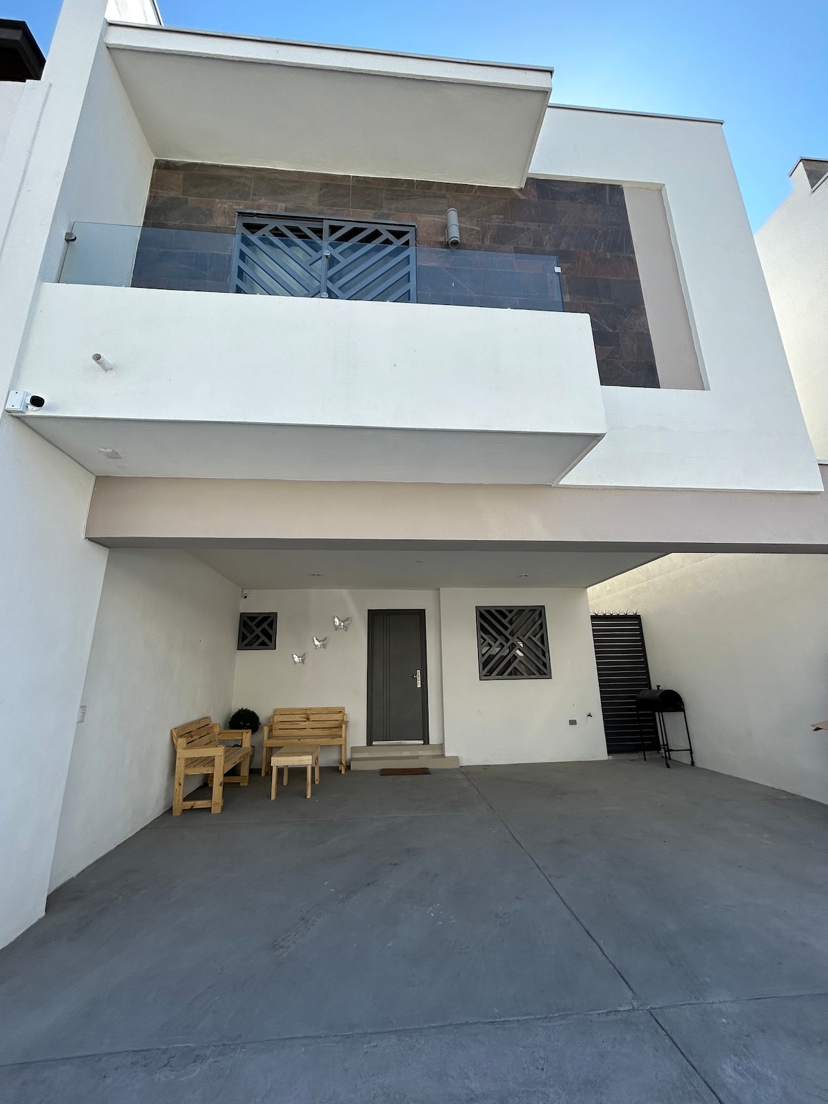 The exterior entrance features a light-colored facade with a modern design. A covered patio provides space for two wooden chairs. The front door is centrally placed, framed by stone accents, and offers access to the welcoming interior.
