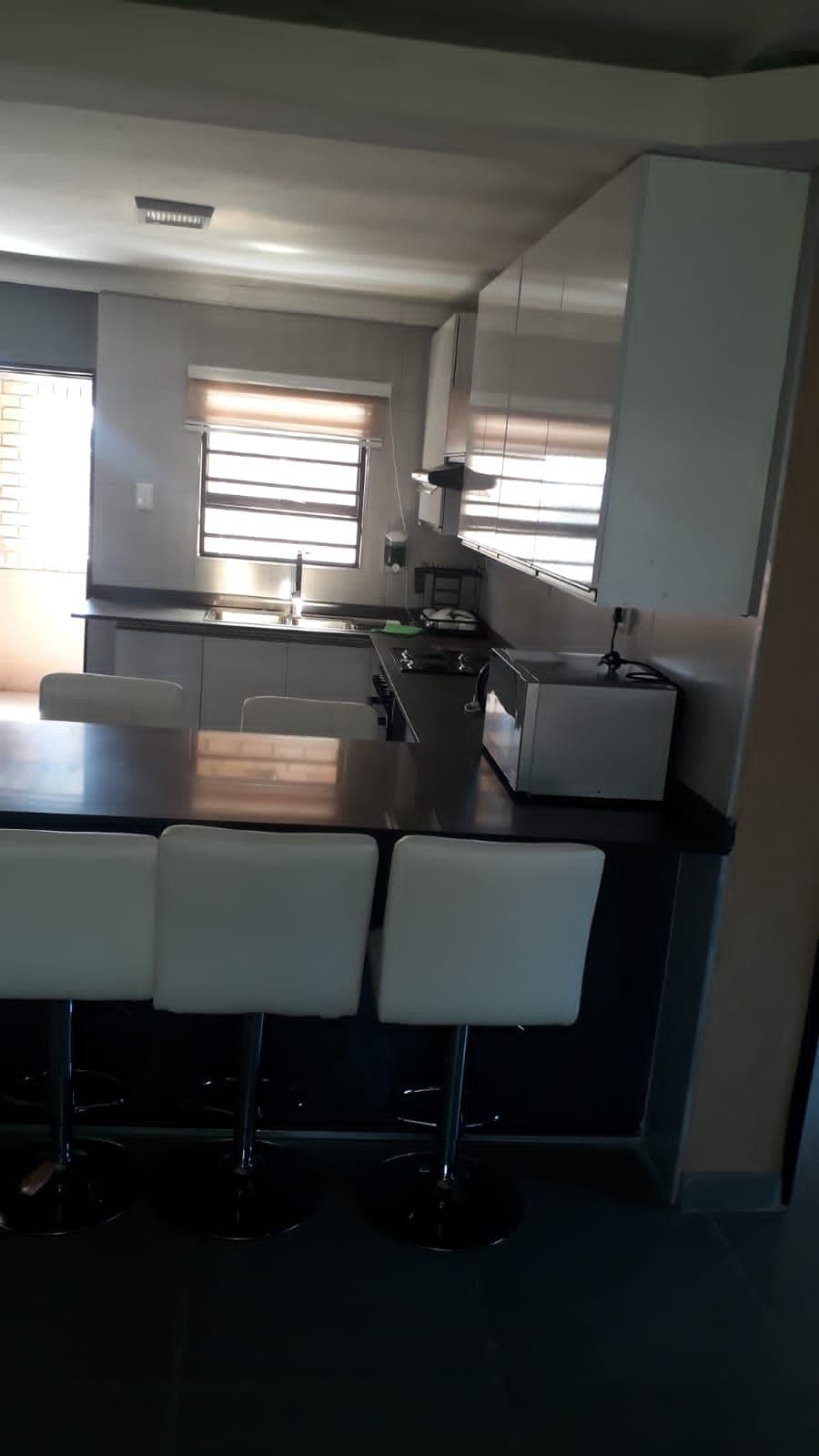 A modern kitchen is presented with sleek cabinetry and a large countertop. Bar stools with white upholstery are arranged around the counter. Natural light streams in through the windows, highlighting the spacious layout and contemporary appliances.