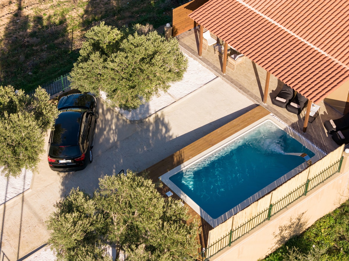 An aerial view of the outdoor space reveals a private swimming pool beside a wooden deck. A black car is parked nearby under the shade of olive trees. The surrounding area is bordered by a fence, enhancing the sense of privacy.
