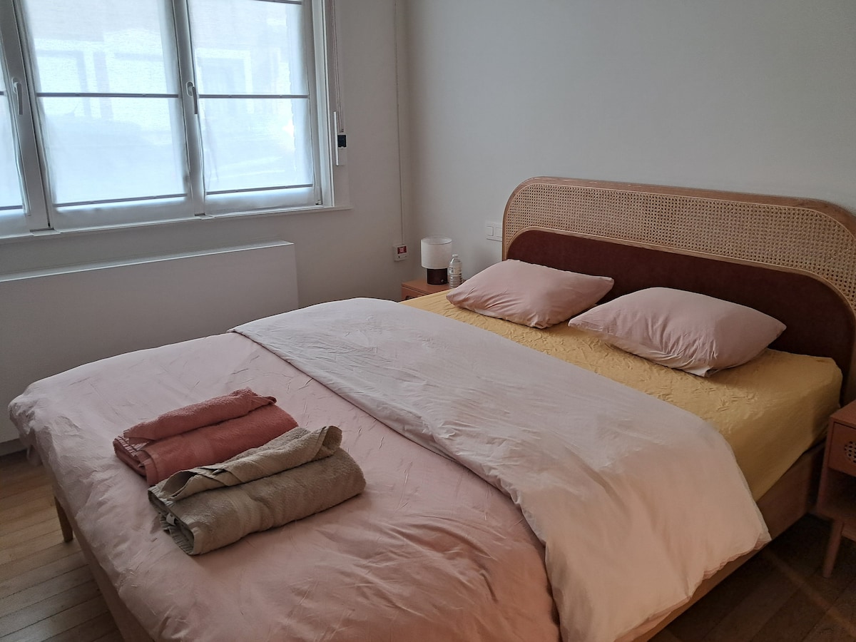 A bright bedroom features a spacious bed with neutral-toned bedding and two pillows. Soft textiles are neatly arranged at the foot of the bed. Natural light fills the room through large windows, creating an airy ambiance.