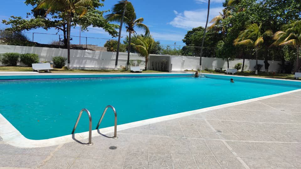 A large swimming pool is surrounded by neatly landscaped gardens. Palm trees are visible around the perimeter, providing shade. The clear turquoise water reflects the blue sky, while sun loungers are positioned along the deck for relaxation.