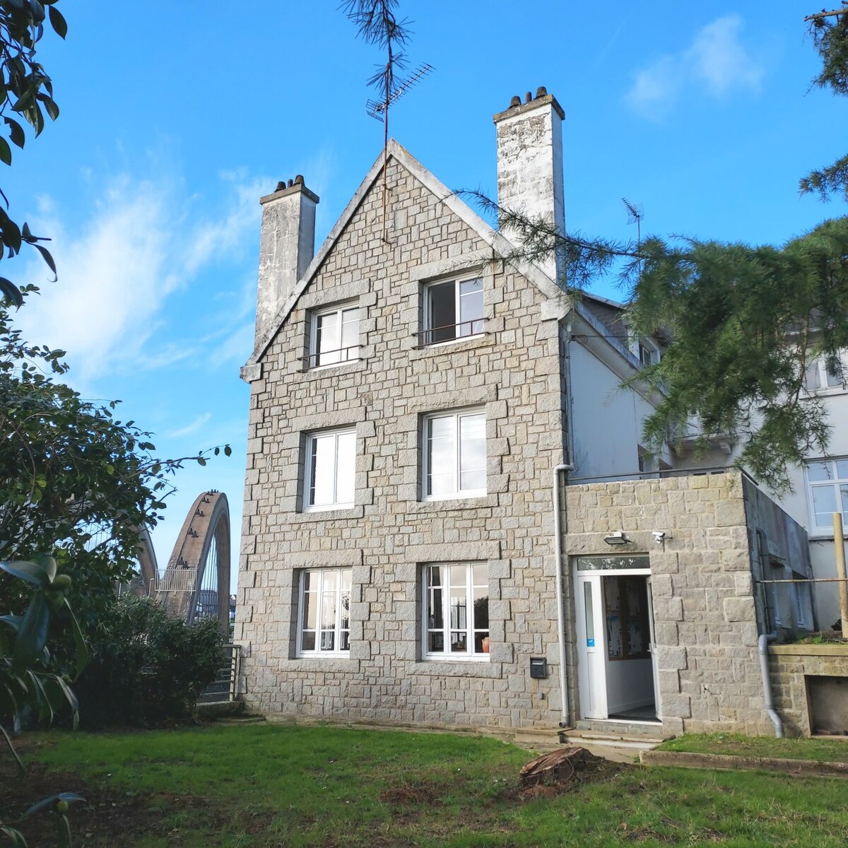 The exterior of the house features a stone façade with multiple windows, revealing a harmonious blend of architecture and nature. A large arching bridge is visible in the background beneath a clear blue sky. The surrounding area includes a grassy space, enhancing the outdoor appeal.