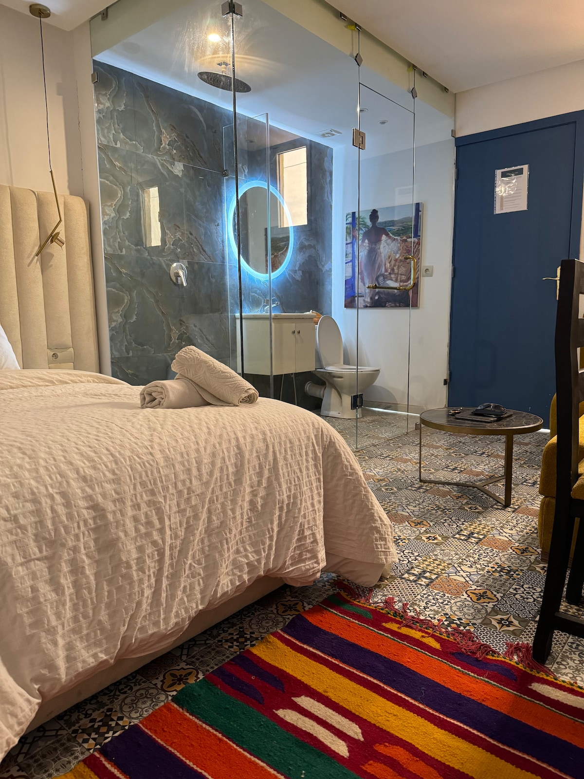 A cozy sleeping area is highlighted by a light-colored bedspread and neatly arranged towels. A modern glass wall reveals a stylish bathroom with a circular mirror. Colorful floor tiles and a vibrant area rug enhance the space, complemented by a small coffee table and a chair.