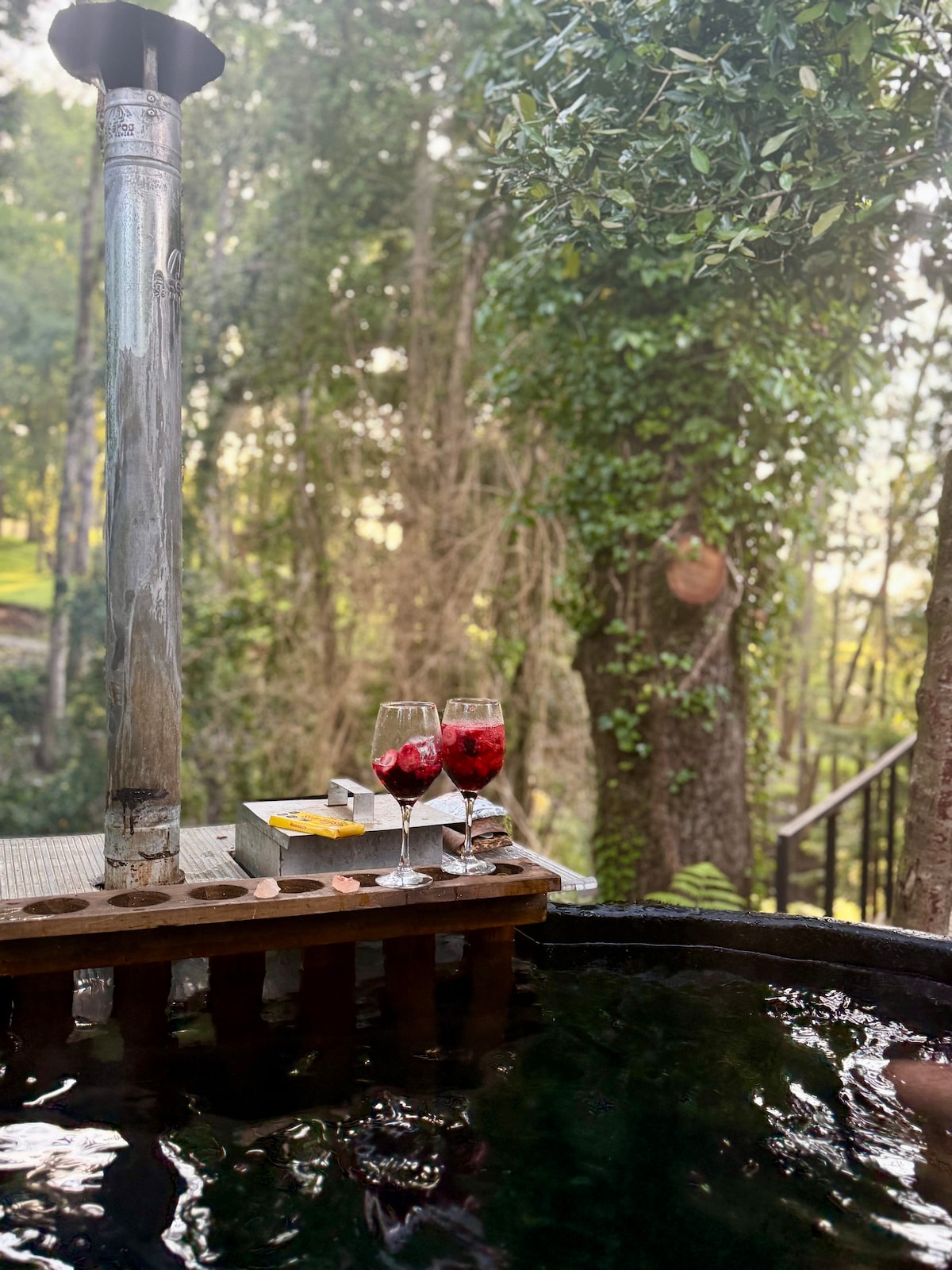 Two glasses filled with a red beverage are placed on a wooden ledge next to an outdoor hot tub. Lush greenery surrounds the area, and sunlight filters through the trees, creating a serene atmosphere.