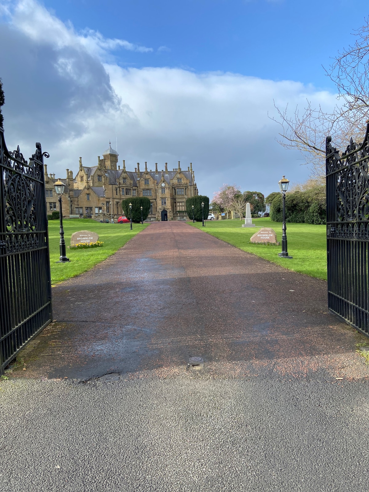 A grand entrance is visible through ornate iron gates, revealing a long paved driveway leading to an impressive historical building. Lush greenery and well-maintained lawns frame the path, while decorative lampposts and sculptural elements add character to the picturesque landscape.