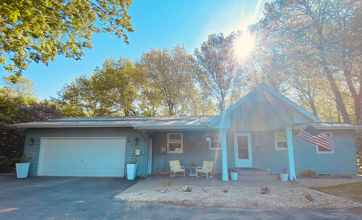 A charming single-story house is set against a bright, sunny sky. The exterior features light blue siding, a spacious driveway, and a neatly landscaped entrance, complete with potted plants and a welcoming flag. Two white rocking chairs are positioned on the front porch.