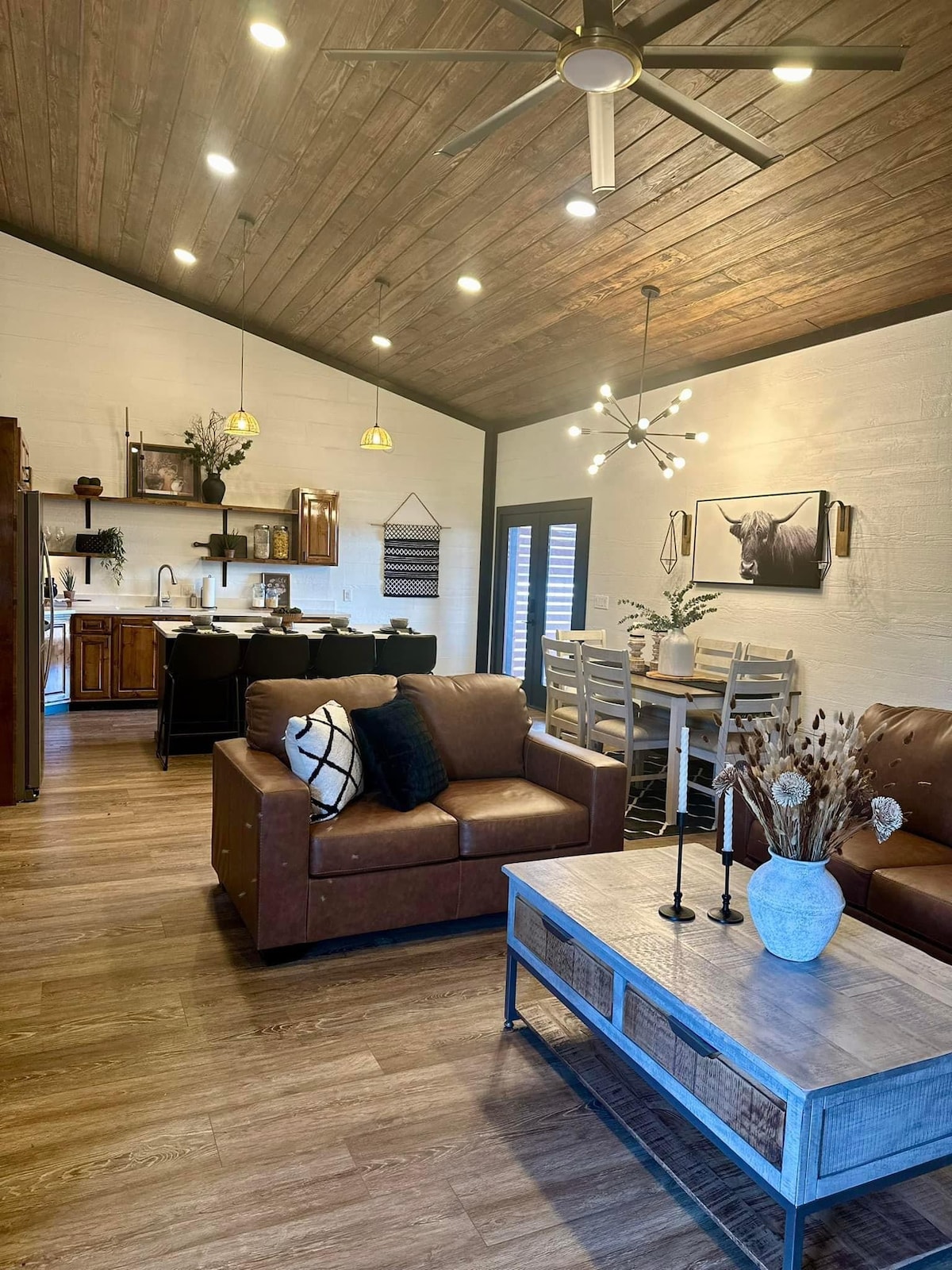 A spacious living area features a brown leather sofa and a wooden coffee table. The dining space includes a light wood table surrounded by chairs. Natural light is invited in through the window and a decorative wall piece is displayed. The ceiling showcases wooden beams with modern light fixtures.