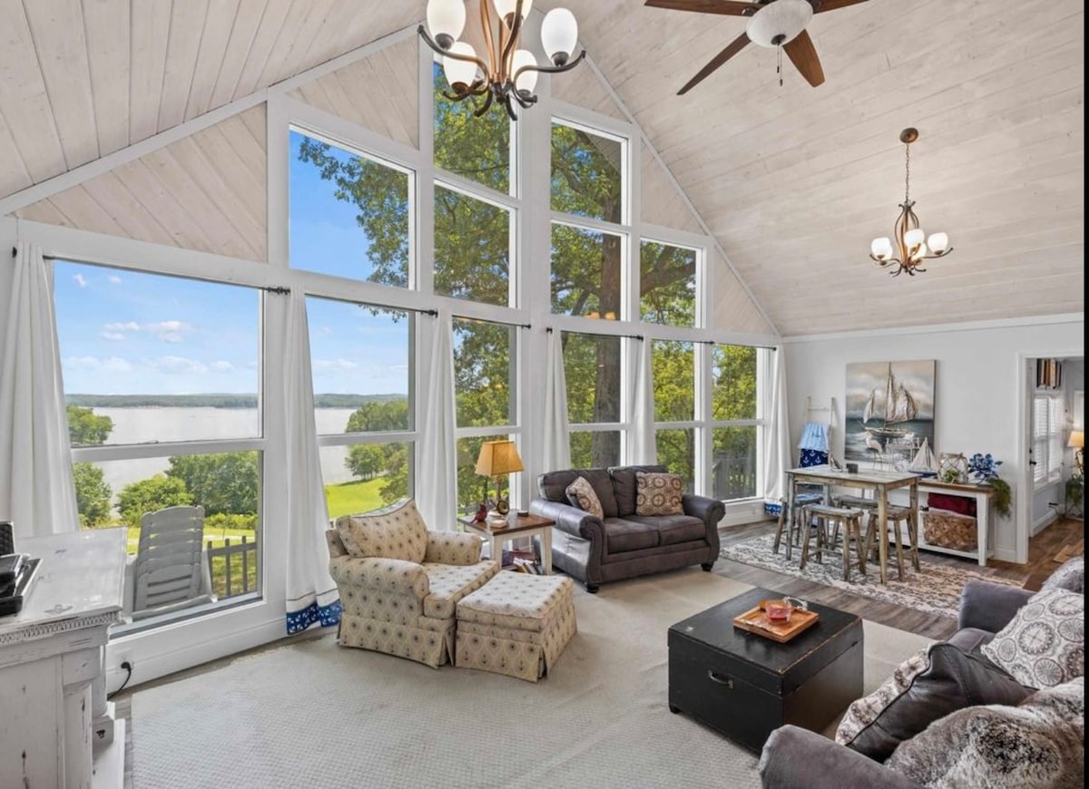 A spacious living area features large windows offering views of the surrounding lake and greenery. Cozy seating includes armchairs and a sofa, complemented by a decorative coffee table. Natural light fills the room, highlighting the wood-paneled ceiling and stylish fixtures.