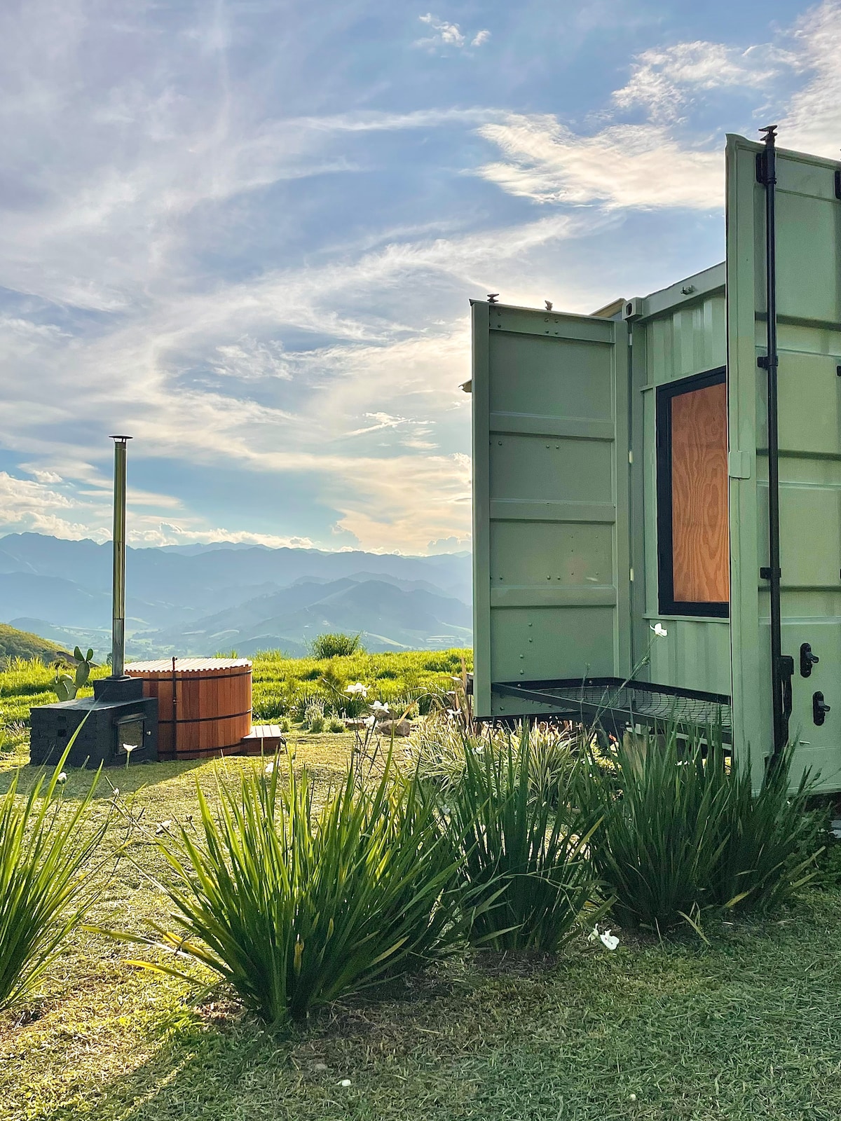 A view of the exterior of a shipping container-style structure is displayed, with a wood-fired hot tub nearby. Lush grass and vibrant greenery surround the area, while mountains are visible in the distance under a partly cloudy sky.