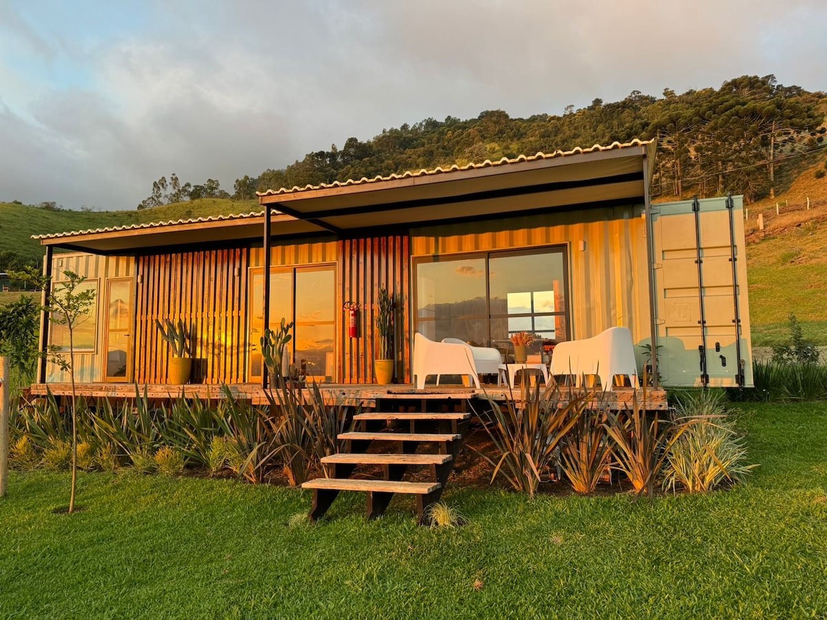 A modern structure created from shipping containers is situated on a green lawn, surrounded by native plants. A wooden deck leads to spacious windows that offer views of the landscape. The design features a covered area with outdoor seating, illuminated by warm sunset light.