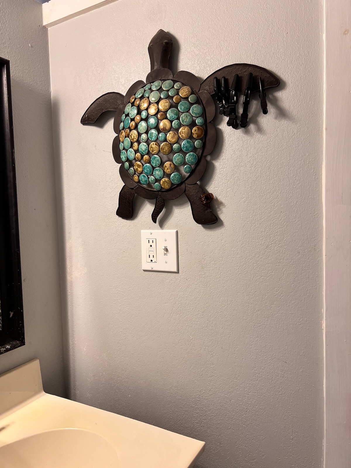 A decorative turtle wall piece is mounted on a muted gray wall, featuring turquoise and gold accents. Nearby, a white sink with minimalistic design sits, complemented by a black-framed mirror reflecting the room's subtle colors. Emphasis is placed on the turtle's intricate design and the wall’s texture.