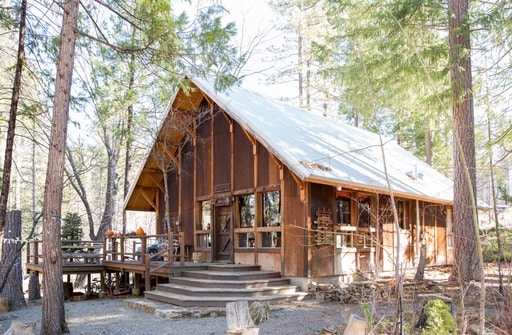 A spacious cabin is nestled among tall trees, featuring a sloped roof and large windows that invite natural light. A wooden deck with steps leads to the entrance, surrounded by peaceful greenery, creating a calming atmosphere for relaxation.
