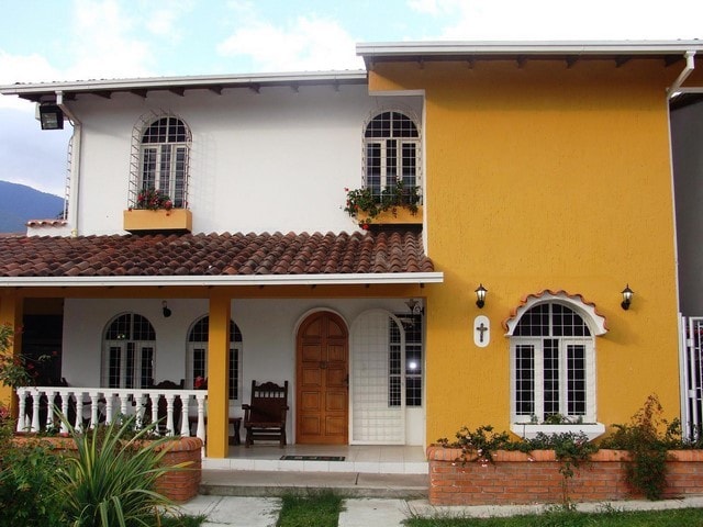 The two-story house features a vibrant yellow exterior complemented by white window frames and decorative ironwork. A welcoming arched wooden door is centered beneath a balcony adorned with potted plants. The front porch is lined with a low brick wall and includes a wooden chair.