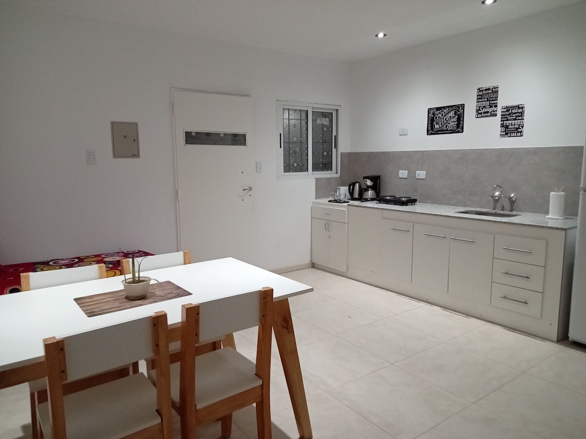 A modern kitchen is accompanied by a dining area. The kitchen features sleek cabinetry, a stove, and a countertop with appliances. A simple dining table with light-colored chairs is positioned nearby, creating a functional space that is bright and inviting.