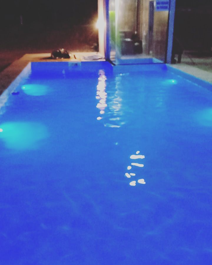 A clear swimming pool is illuminated by soft blue lights, creating a tranquil ambiance at night. The pool's shimmering surface reflects the surrounding environment, with a modern structure visible in the background.