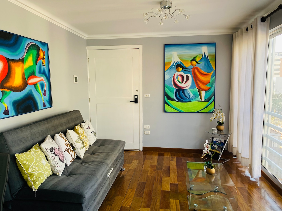 A modern living area features a sleek gray sofa adorned with decorative pillows. On the walls, two vibrant pieces of artwork bring color and character. Natural light enters through a large window, illuminating the space. A glass coffee table complements the warm wooden flooring.