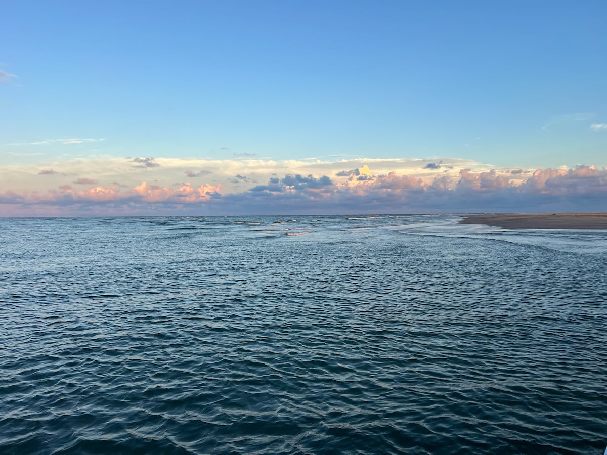 The calm waters reflect soft hues of blue and pink from the sky during twilight. Gentle waves create a textured surface, stretching toward a sandy shore in the background. Fluffy clouds add dimension to the serene coastal landscape.