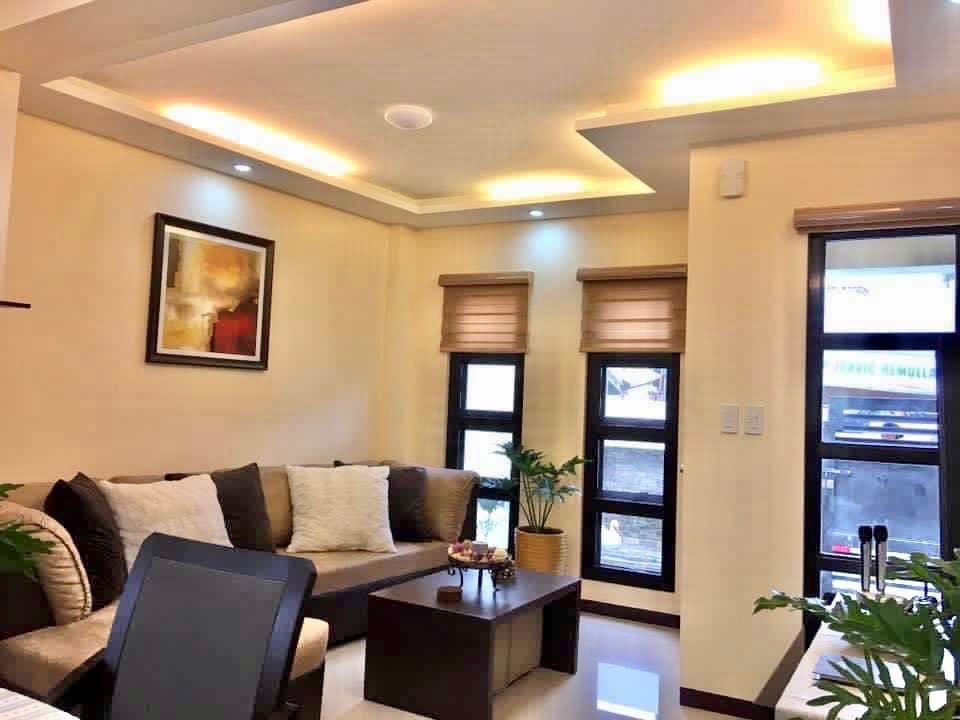 A cozy living area is presented with a comfortable sectional sofa adorned with decorative pillows. A small wooden coffee table sits at the center, displaying a modest arrangement of plants. Natural light enters through two windows fitted with blinds, illuminating the softly colored walls.