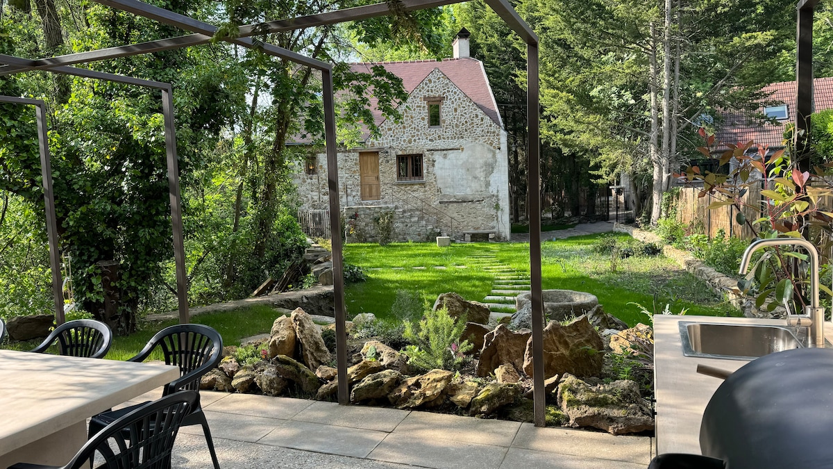The garden area features a stone pathway leading to a lush green lawn surrounded by trees and decorative rocks. A dining table with black chairs is positioned in the foreground, while a charming stone wall house adds historical character in the background.