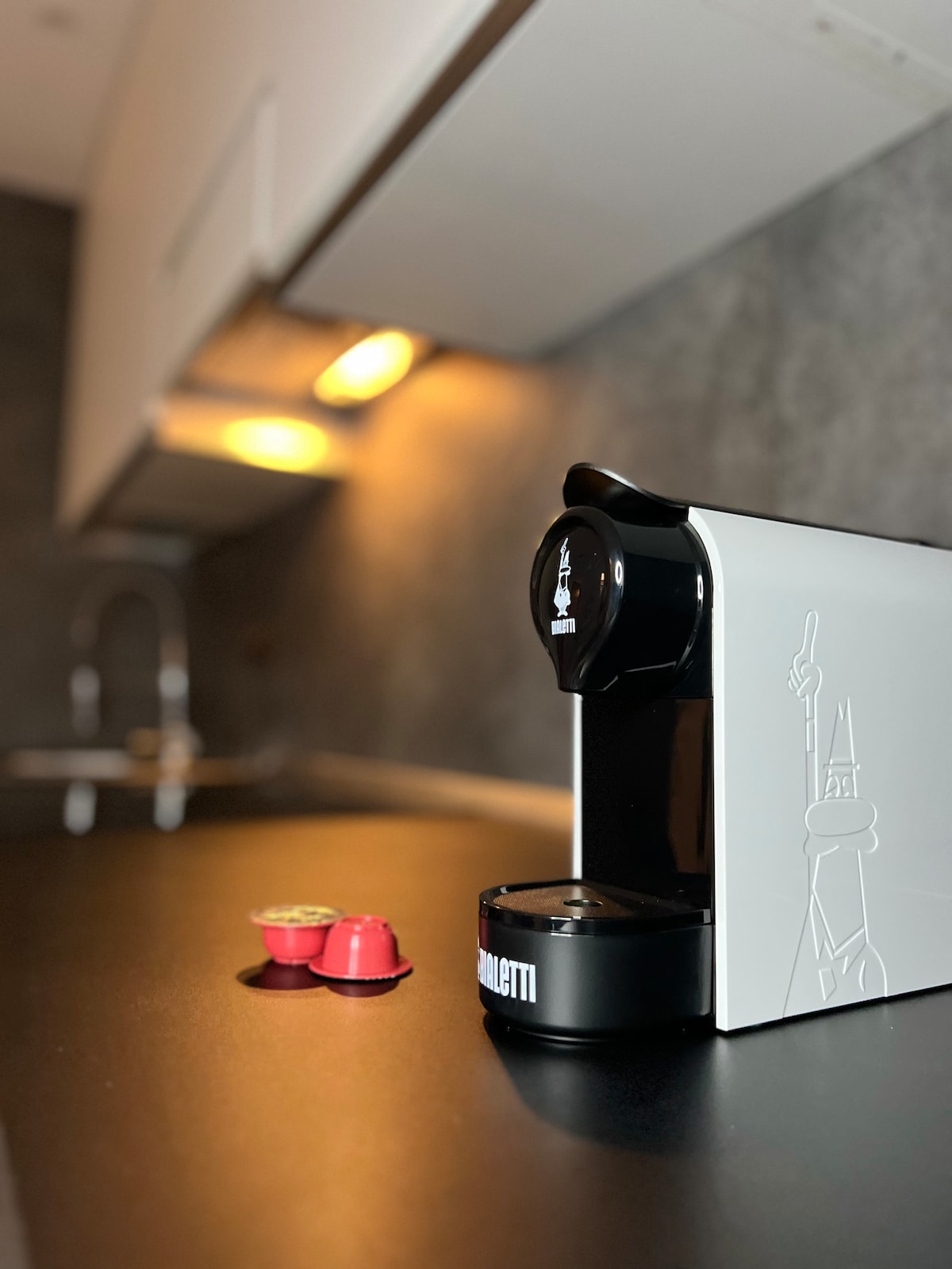 A modern coffee machine sits on a sleek black countertop, accompanied by two colorful coffee capsules. The kitchen features minimalist cabinetry with soft lighting, highlighting the contemporary design.