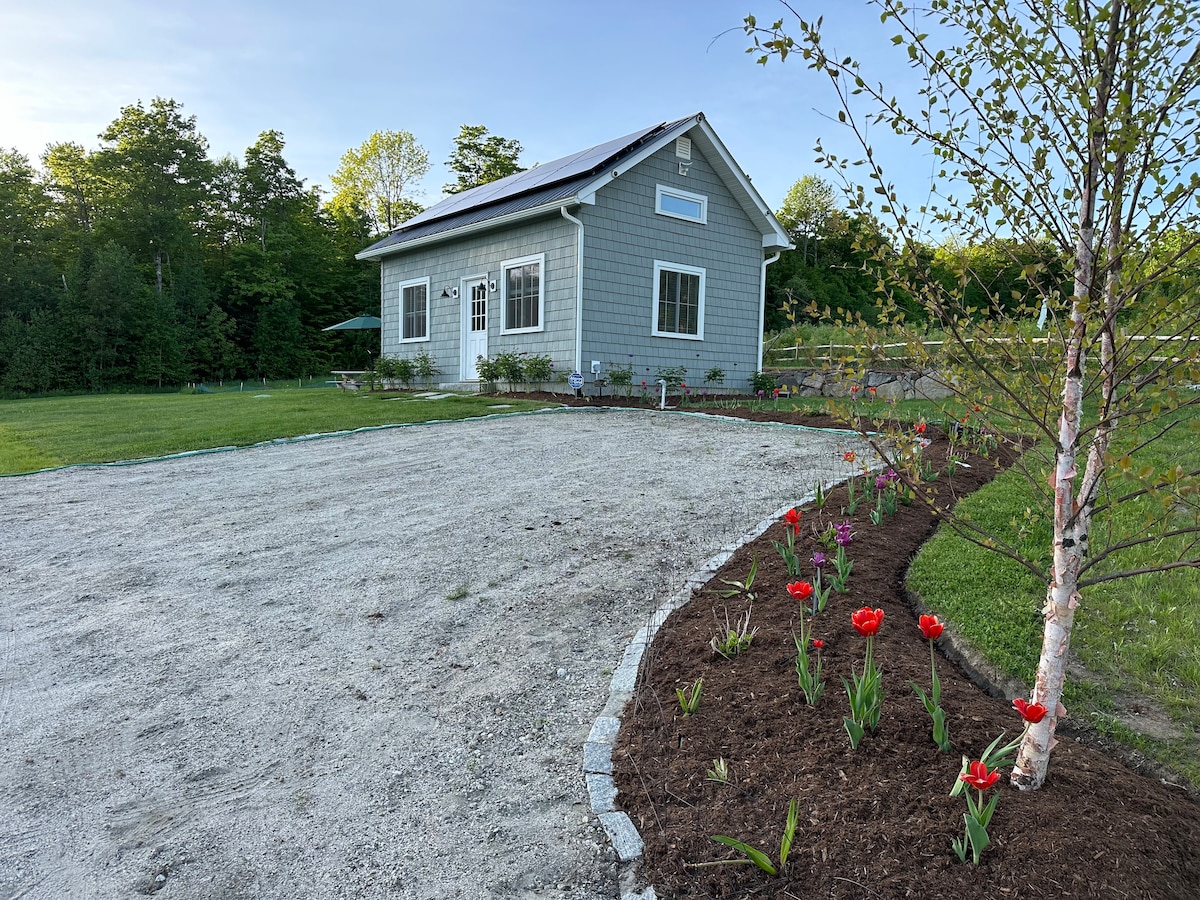 A modern gray house is set within a landscaped area, featuring freshly arranged flower beds with colorful blooms. The surrounding lawn is neatly maintained, and a gravel pathway leads to the entrance. Green trees and a clear sky add to the serene outdoor setting.