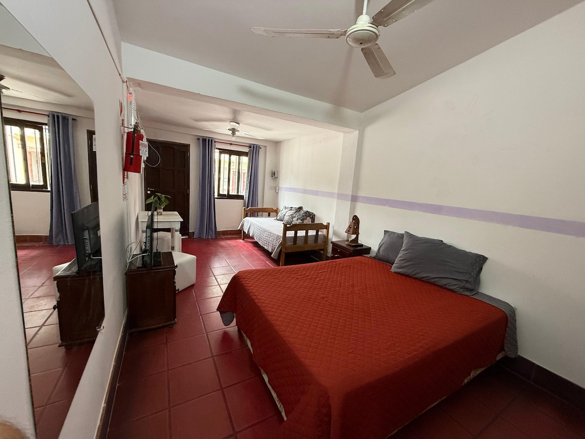 A spacious room is presented with two beds, one covered in a red bedspread and the other with a gray blanket. A television is positioned on a wooden cabinet, and large windows allow natural light to flow in, framed by curtains. A mirror reflects the room's layout.