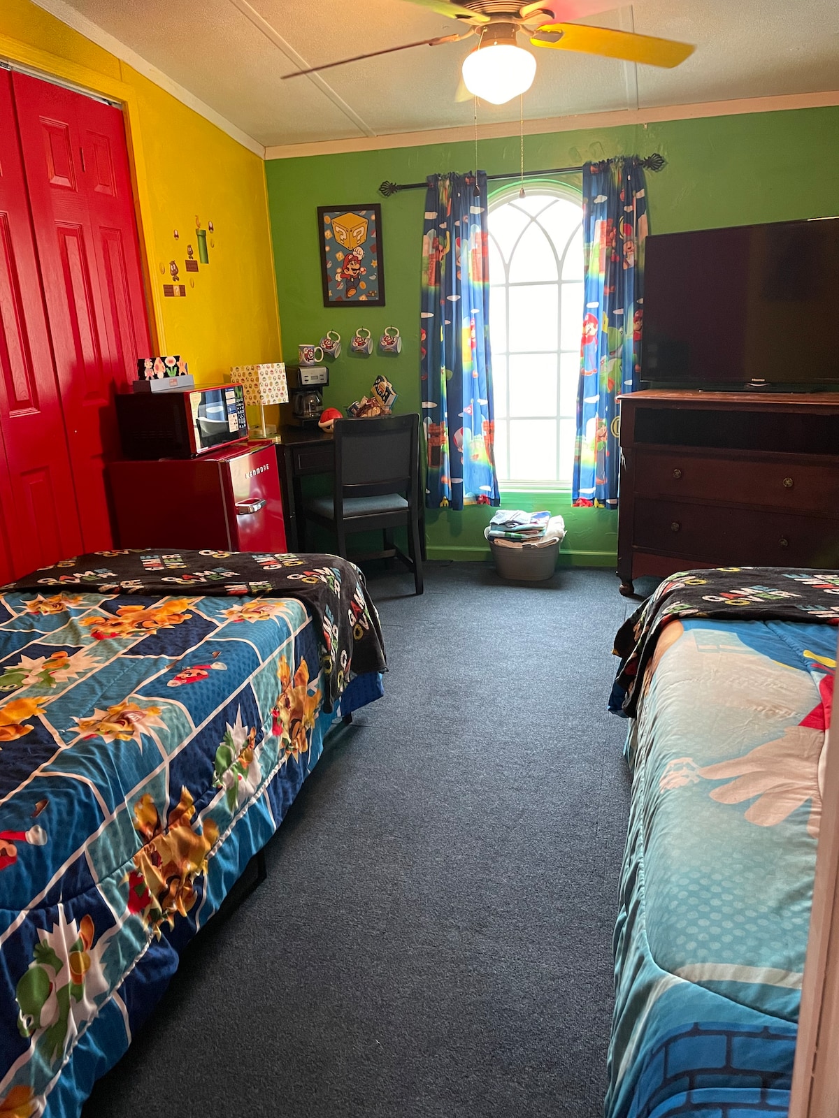 A bright children’s room features two twin beds covered in colorful bedspreads. A small desk and chair are positioned beside a television, offering a playful theme. The walls are painted in vibrant yellow and adorned with cheerful decorations, while a window with patterned curtains allows natural light to enter.