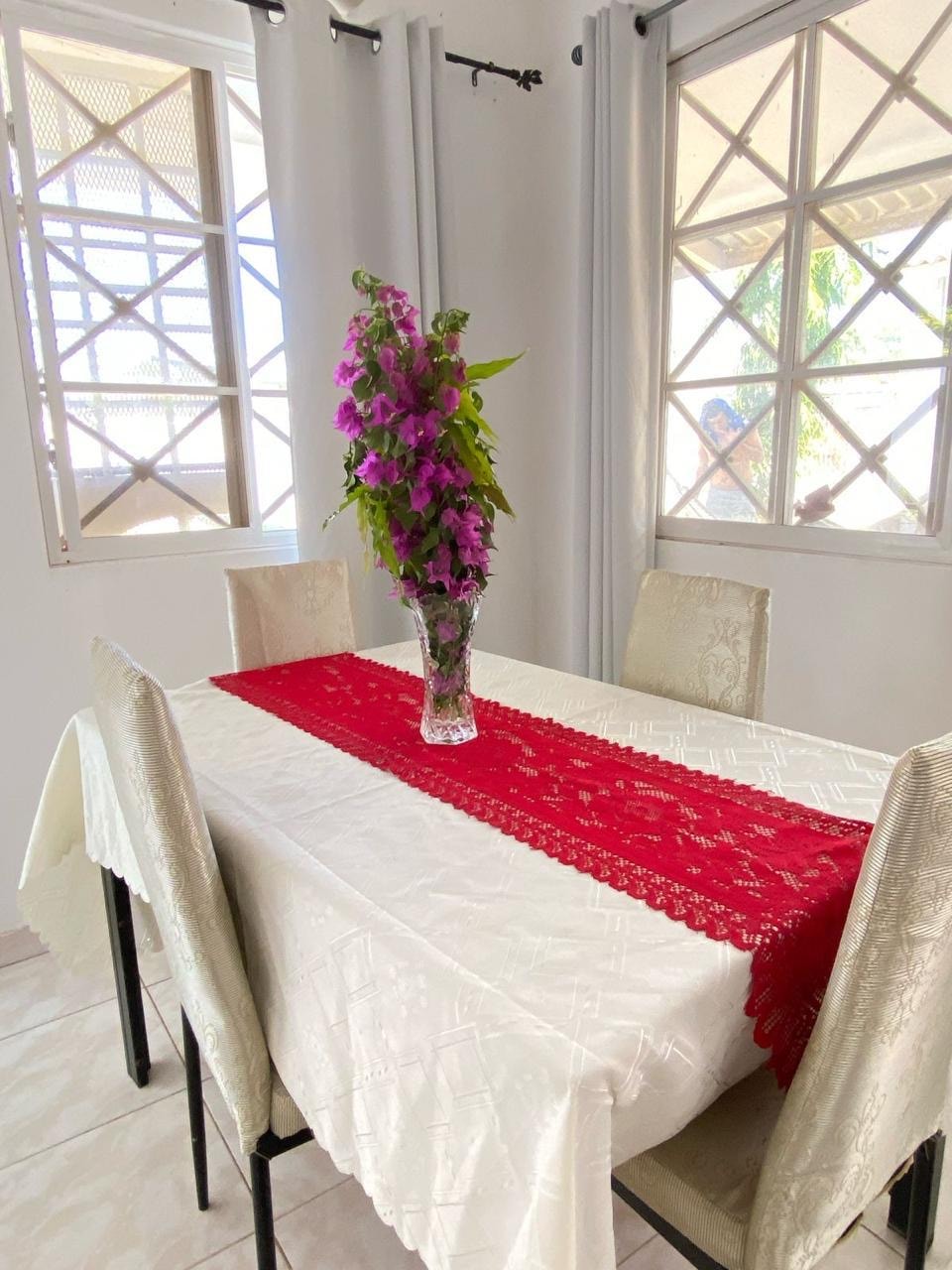A dining area is presented with a simple table covered in a white tablecloth. A vibrant red lace runner adds color, complemented by a clear vase holding fresh purple flowers. Natural light filters in through windows featuring decorative grilles.