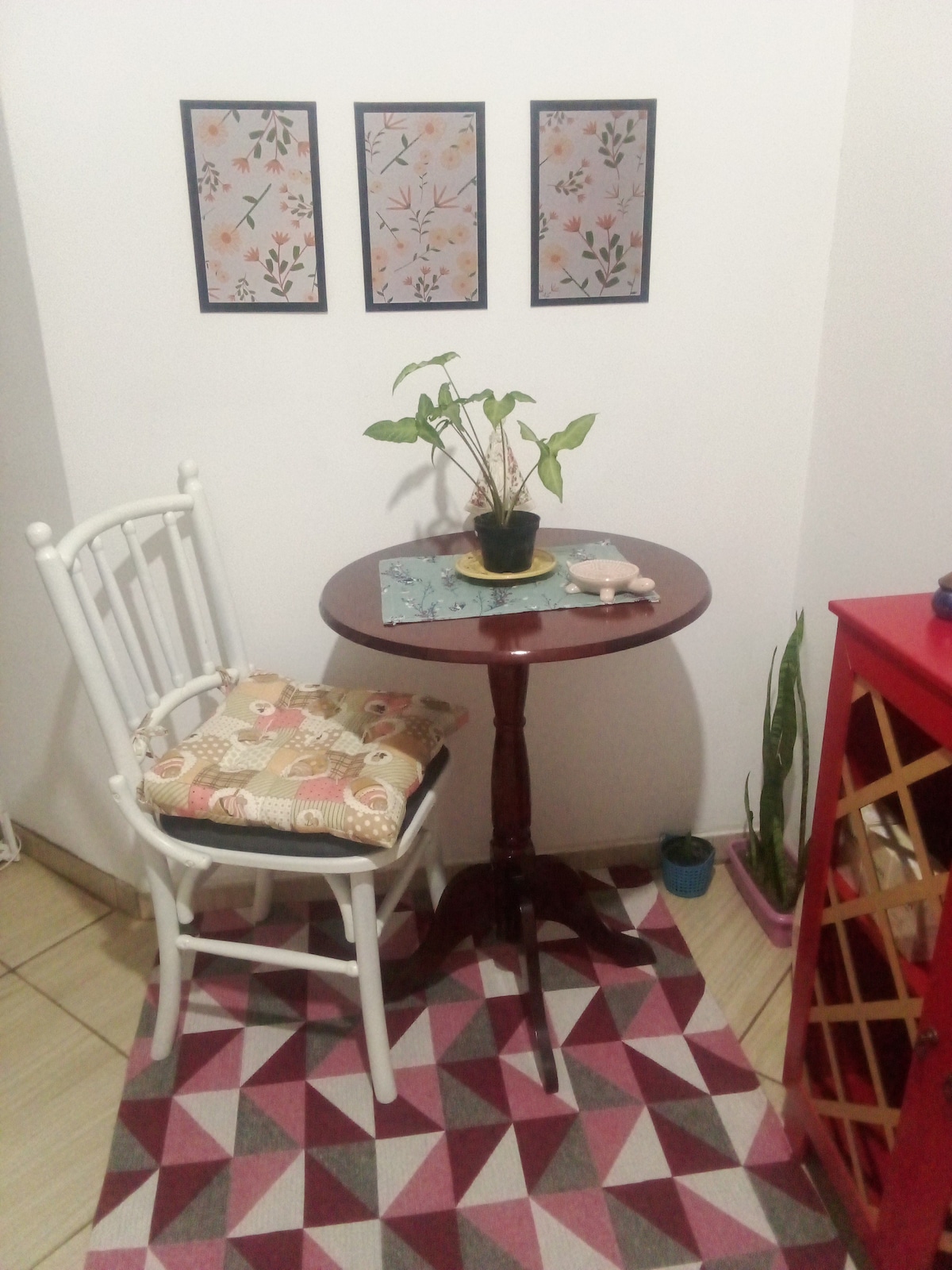 A small dining area features a round wooden table with a green plant in a pot and decorative objects. A white chair with a patterned cushion complements the setting. The wall is adorned with three framed botanical prints, and a colorful geometric rug adds texture to the floor.
