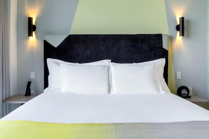 Ëndrra fluturuese mbi çarçafë të freskët, nën ndriçimin elegant dhe modern të kokës së krevatit.