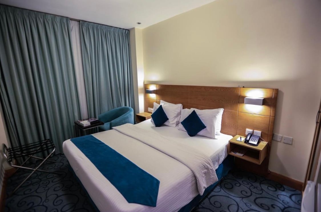 A sleeping area features a spacious bed dressed in white linens with an accent throw. Two decorative pillows are placed at the head. A comfortable chair is situated beside the bed, and floor-to-ceiling curtains frame a window, allowing natural light to illuminate the space.