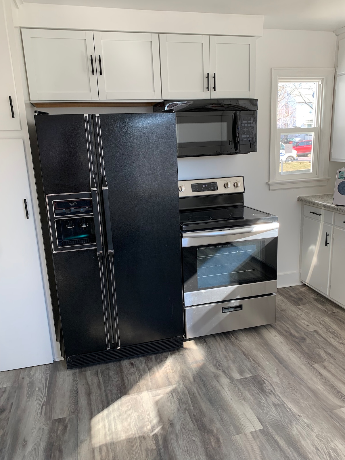 A modern kitchen showcases sleek black appliances, including a refrigerator with double doors and an oven range. Natural light enters through a nearby window, highlighting the clean lines of the cabinetry and the contemporary flooring.