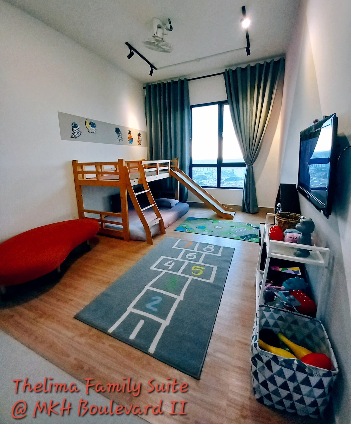 A children's play area is featured, equipped with a wooden bunk bed and a colorful rug displaying a hopscotch design. A small red sofa is positioned nearby, with storage baskets filled with toys located in the corner. Large windows allow natural light to brighten the space.
