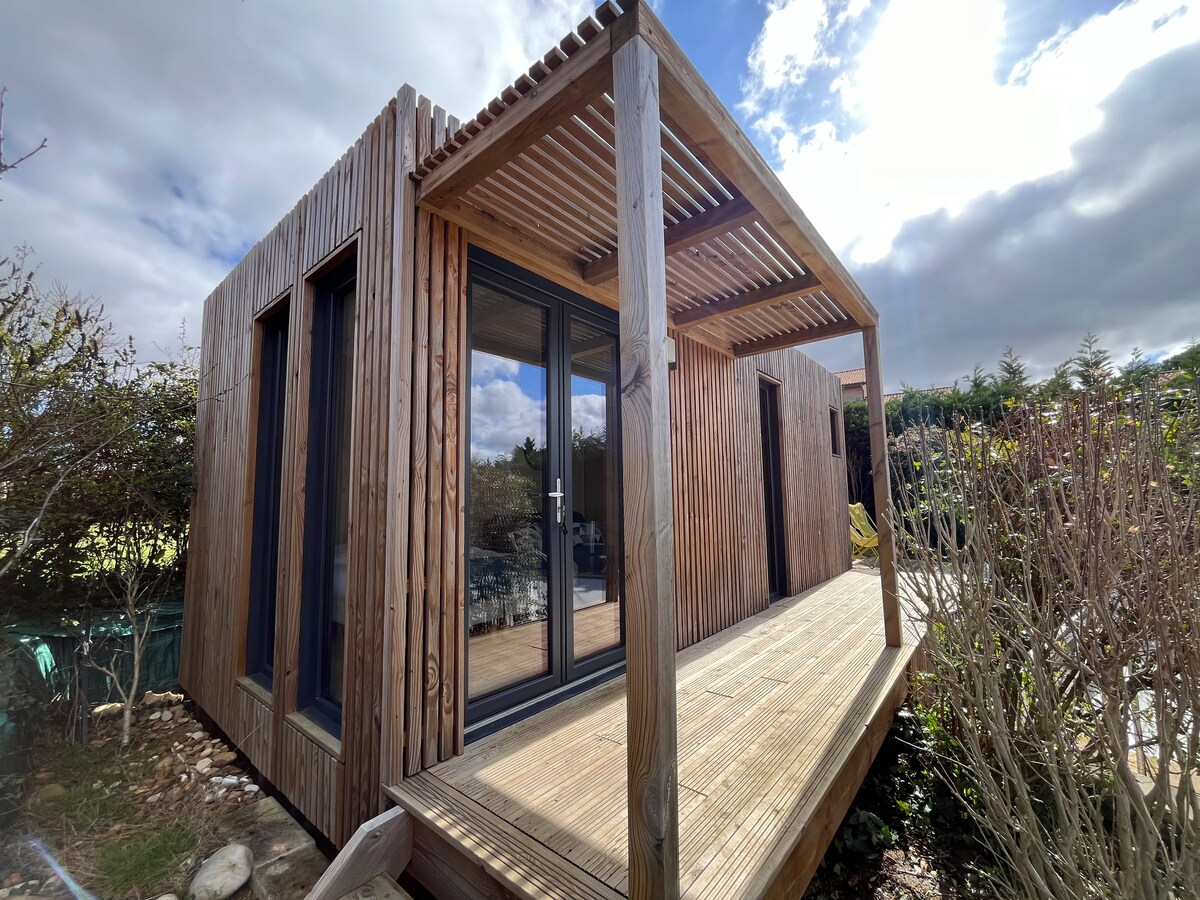 The modern wooden studio features a wraparound deck with a shaded overhang, creating a welcoming entrance. Large glass doors provide views into the interior, while the exterior is framed by greenery and natural landscaping.