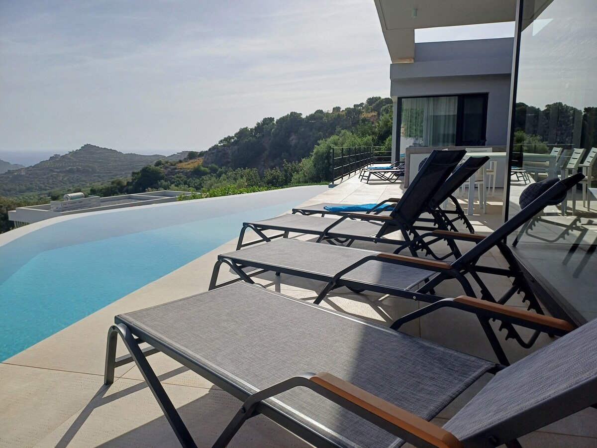 A modern outdoor terrace is presented, featuring several lounge chairs arranged by a serene infinity pool. The pool's water offers reflections of the surrounding hills and sky, while the distant landscape enhances the peaceful atmosphere of this villa.