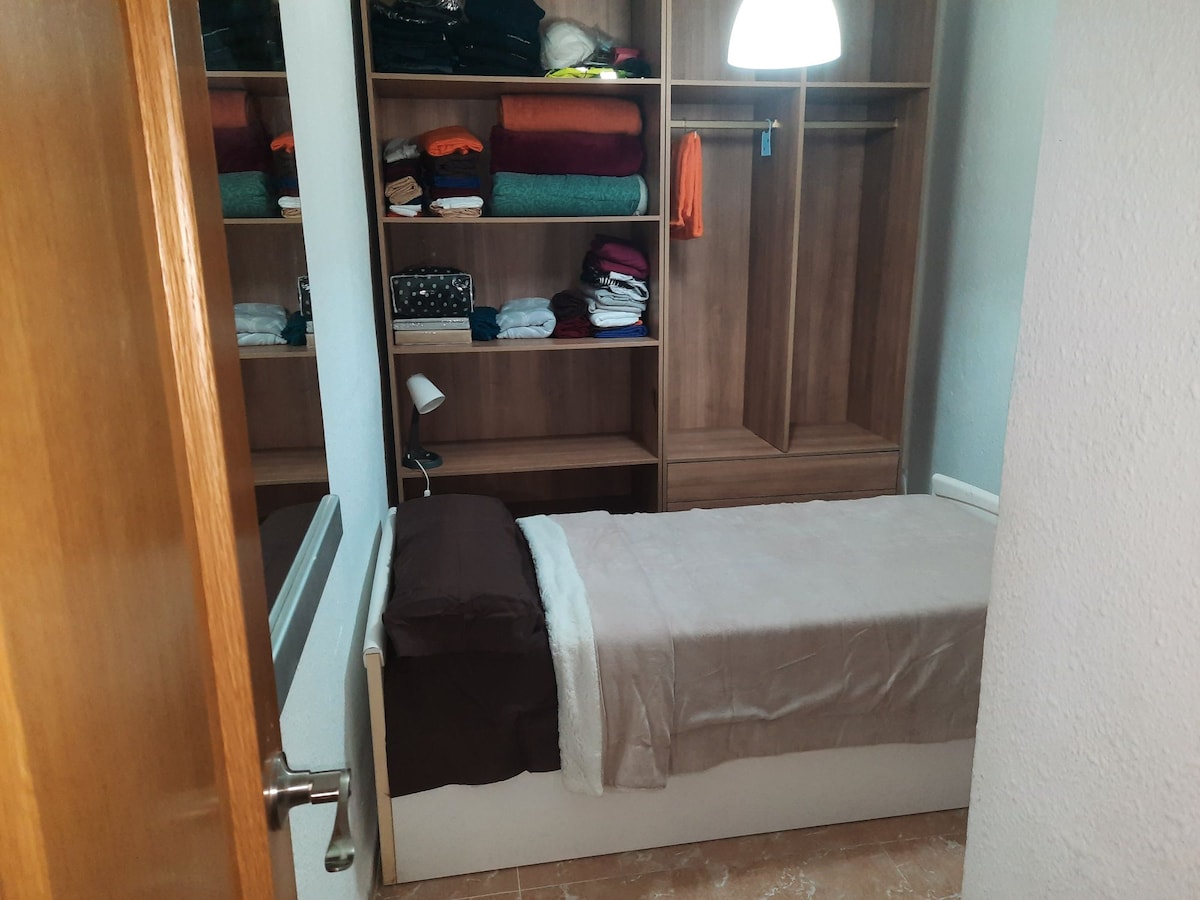 A compact sleeping area is visible, featuring a neatly made single bed with a neutral bedding set. Behind the bed, shelves are filled with folded linens and towels. A small lamp is placed on a bedside table, providing functionality in the space.