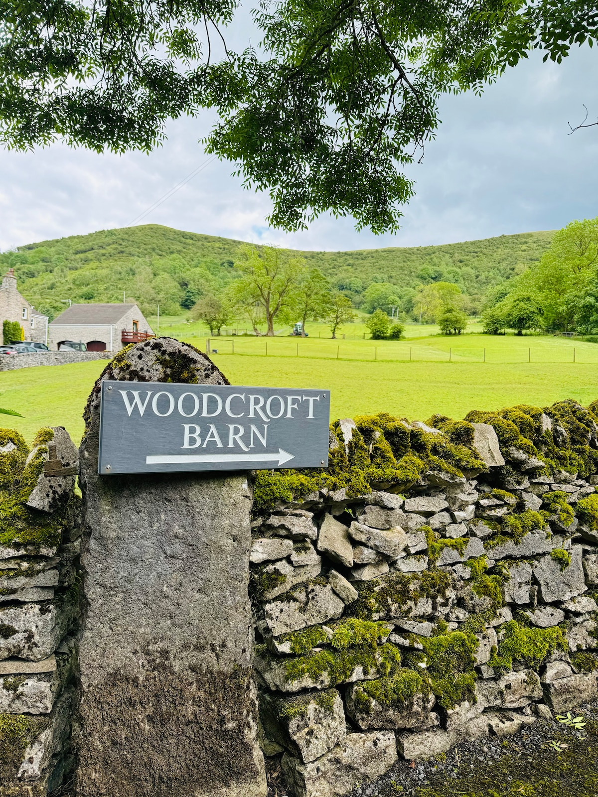 A stone sign labeled 'WOODCROFT BARN' is positioned atop a moss-covered stone wall. Lush green fields extend into the distance, with rolling hills framing the background. A farm building is visible to the left, partially obscured by trees and gentle hill slopes.