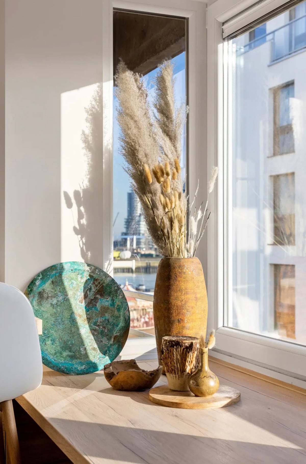 A sunlit windowsill displays an artistic arrangement of decorative items. A tall, textured vase holds dried grasses, while a colorful plate and small sculptures complement the ensemble. Light reflects off the surfaces, creating a serene and stylish atmosphere.