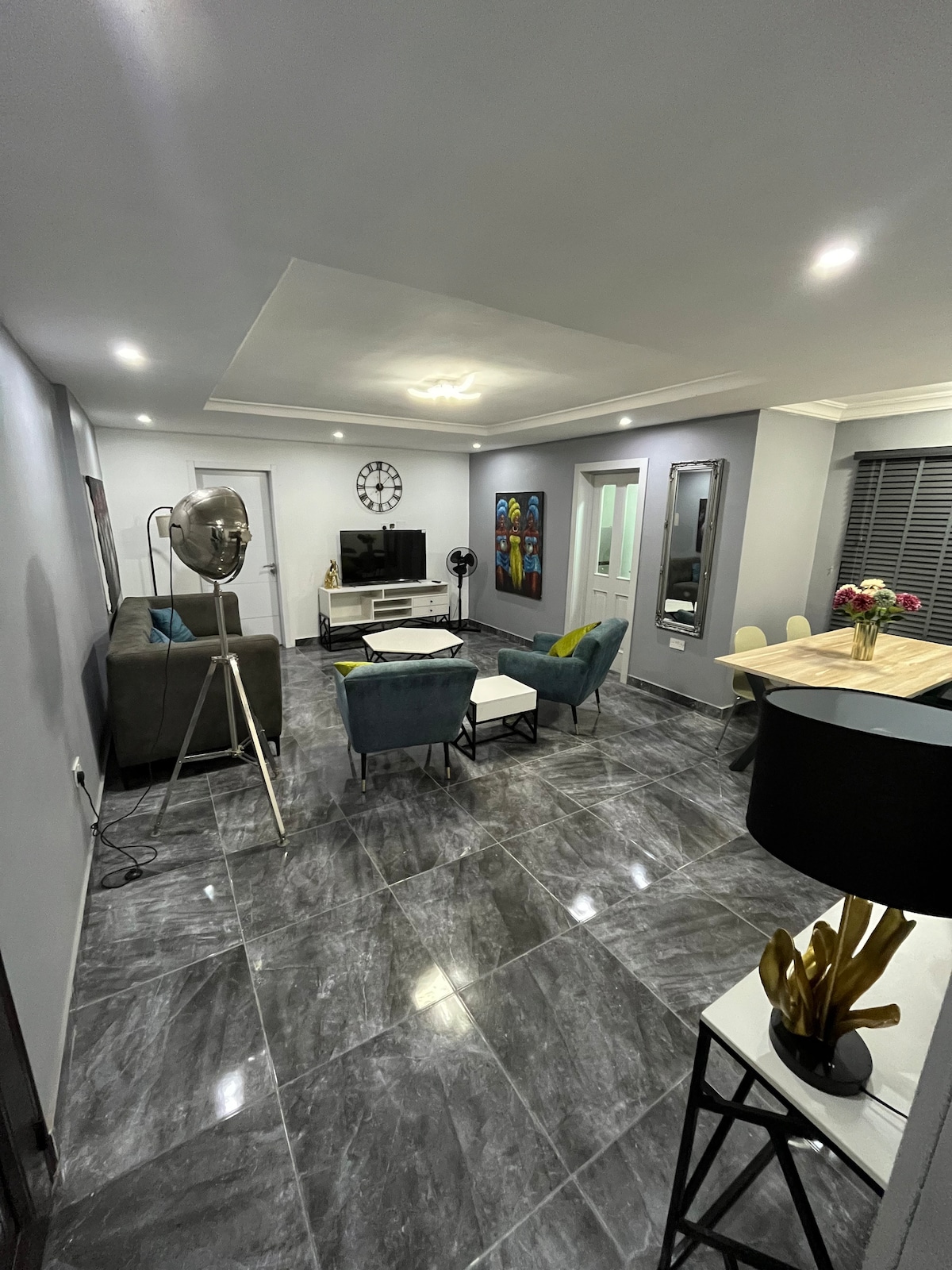 A spacious living area is shown, featuring modern furnishings in calming hues. A sleek entertainment unit is situated against a wall, accompanied by a decorative clock. Soft lighting enhances the ambiance, and a dining area is visible with a wooden table and chairs, adding functionality.