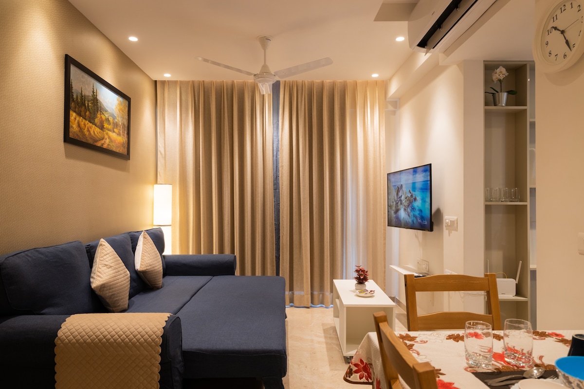 A stylish living area features a dark blue sofa adorned with light-colored pillows. A warm glow is provided by a lamp beside it. Large curtains in a soft hue cover a window, while a wall-mounted TV is visible across from a simple dining table set with glassware.