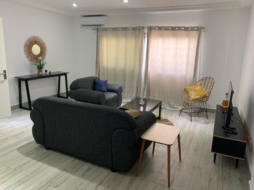A living area is presented with modern furnishings, including a dark gray couch and a round accent chair. Natural light filters through sheer curtains, complementing a small table and a television mounted on the wall. A decorative wall piece adds a touch of style.