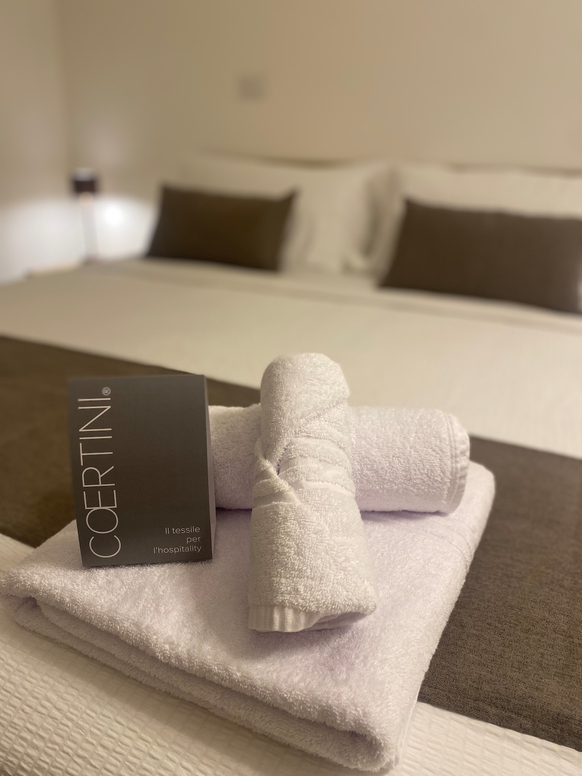A spacious bed is dressed in neutral tones, featuring soft white linens and plush pillows. Fresh, neatly rolled towels are placed on the bed. A hospitality booklet is positioned on the side, offering information about the stay.