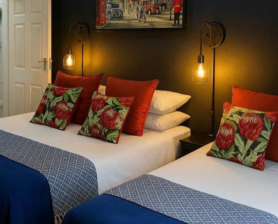 The sleeping area features two spacious beds, adorned with vibrant floral cushions. Each bed is accompanied by nightstands with softly glowing pendant lights. The dark accent wall adds depth, while a piece of art depicting urban life hangs above the beds.