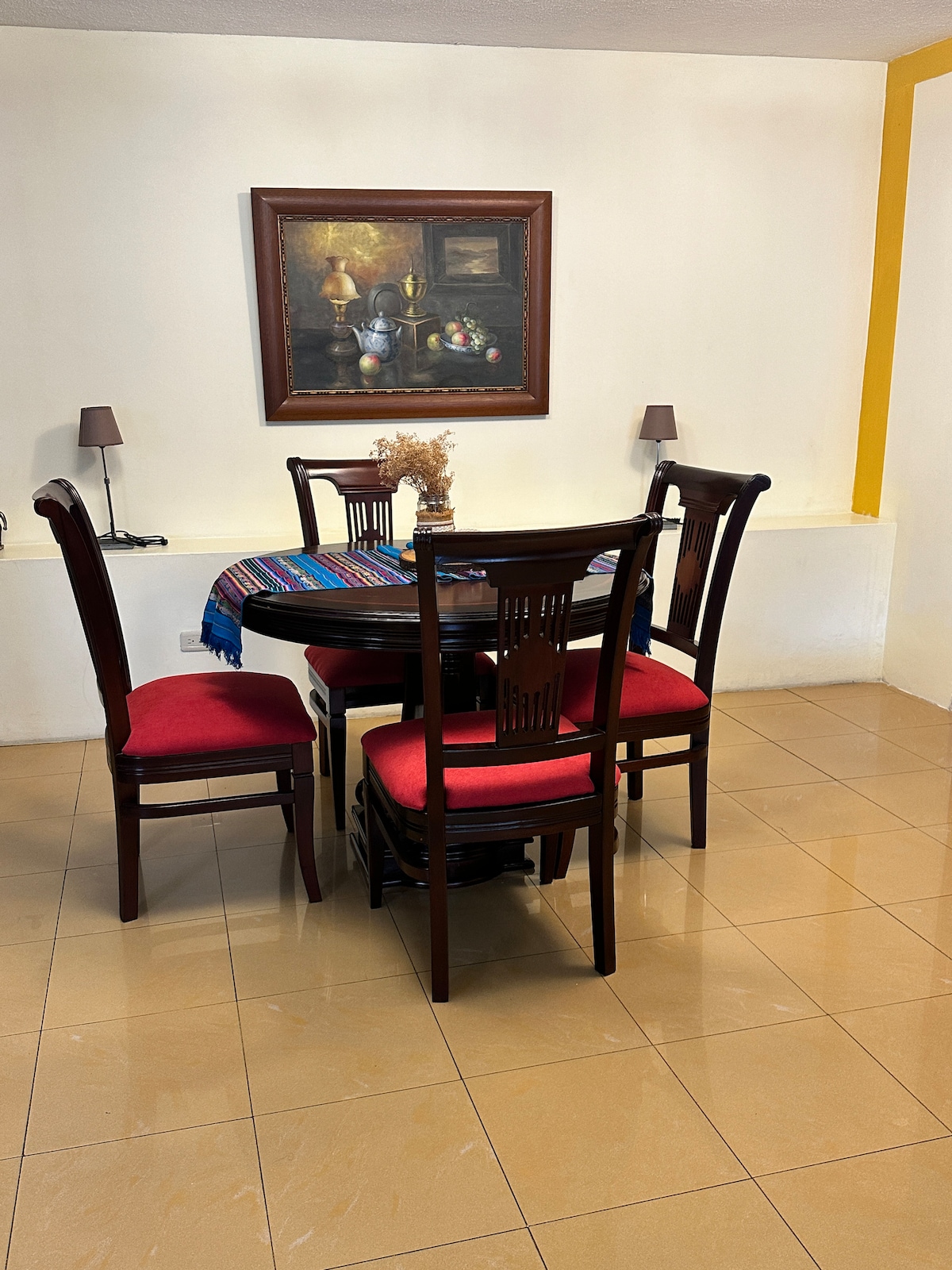 A dining area is presented, featuring a round table surrounded by four wooden chairs with red cushions. Above the table, a framed painting adds character to the space. Soft lighting is provided by two tabletop lamps on either side.